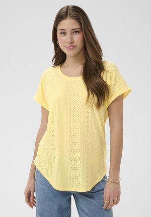 Junge Frau mit langen braunen Haaren, die ein gelbes, kurzärmeliges, perforiertes Oberteil und hellblaue Jeans trägt, vor einem einfarbigen Hintergrund steht.