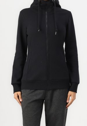Femme portant un sweat à capuche noir zippé avec cordons et un pantalon de jogging gris foncé, debout devant un fond blanc uni.