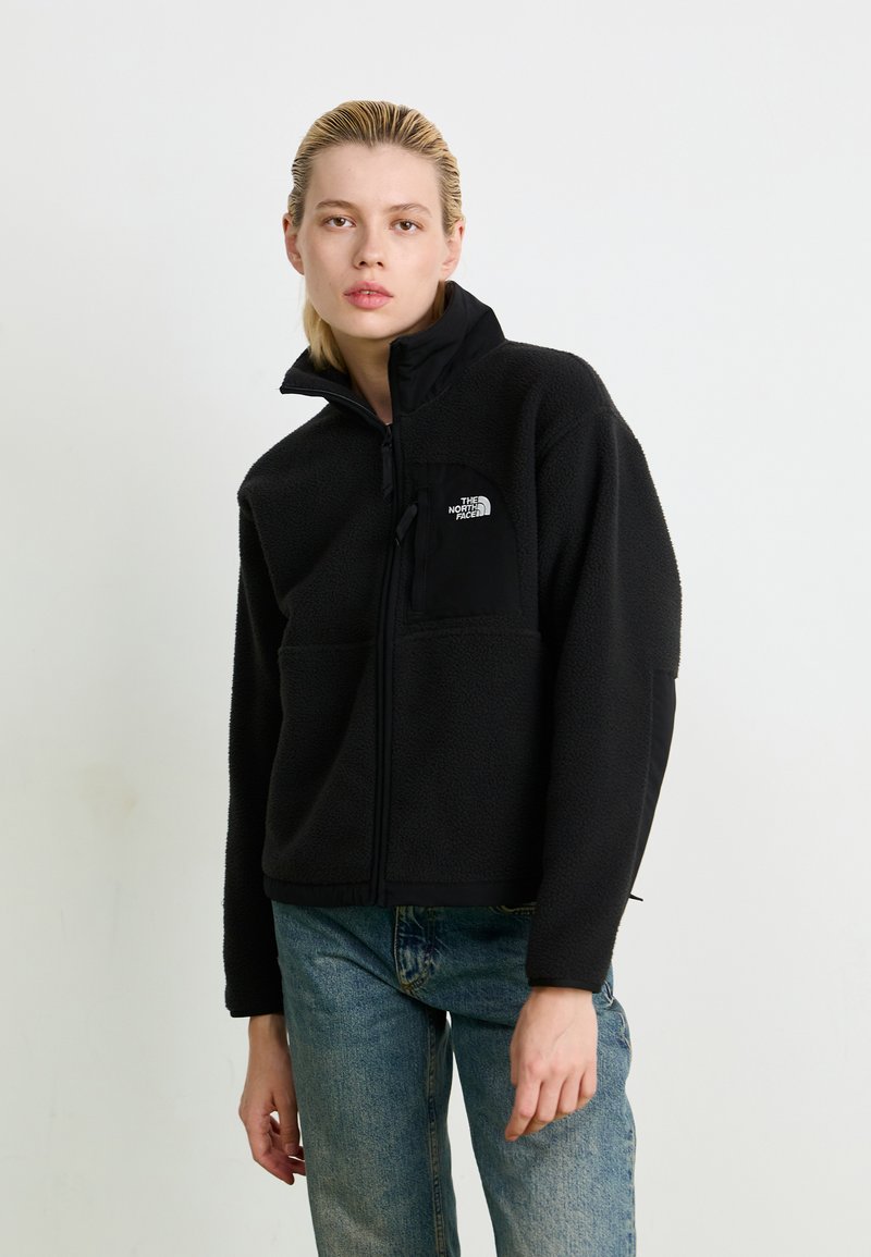 Black fleece jacket with high collar, front zip, two side pockets, and a chest pocket featuring a white logo on the left side.