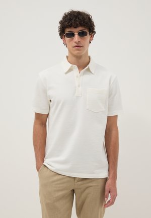 Man met krullend haar, donkere zonnebril, wit gestructureerd poloshirt met een borstzakje en beige broek, staand met een hand in zijn broekzak.