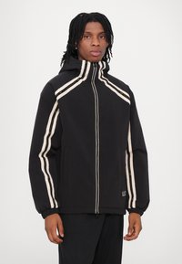 Veste noire avec un col haut, présentant des bandes blanches et dorées le long des manches et une fermeture éclair à l'avant. Tissu texturé.