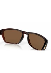 Occhiali da sole color marrone con una montatura spessa in tartaruga scura, dal design angolare e con un accento dorato sulle tempie.