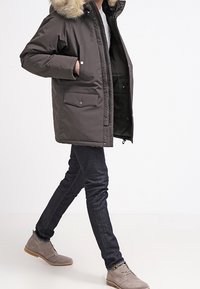 Parka marrone scuro con cappuccio bordato di pelliccia, dotata di chiusura a bottoni a pressione, due tasche frontali e un tessuto liscio e isolante. Indossata con jeans scuri e stivali.