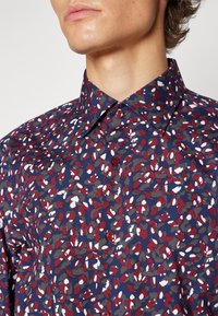 Homme portant une chemise bleu foncé boutonnée avec col, affichant un motif abstrait rouge, blanc et gris, visible du cou à la poitrine.