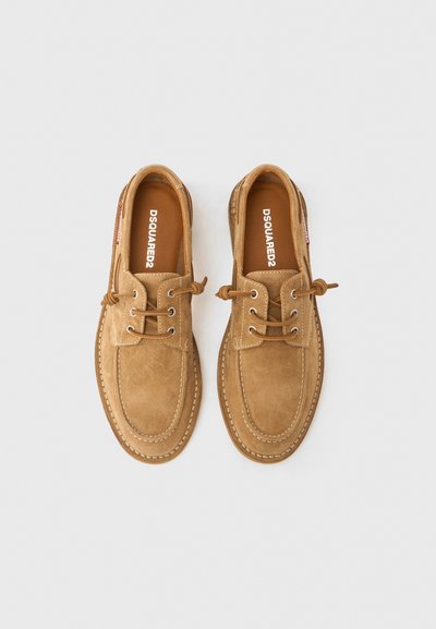 Καφέ παπούτσια ιστιοπλοΐας από σουέτ με στρογγυλερή μύτη, ραμμένες λεπτομέρειες και σχεδιασμό με κορδόνια. Εσωτερικά διαθέτουν το λογότυπο του εμπορικού σήματος σε λευκό πάνω σε μπεζ πάτο.