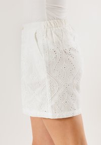 Pantaloni corti bianchi ricamati con un design a fantasia e aperture ad occhio. Vita elasticizzata per comfort e vestibilità rilassata.