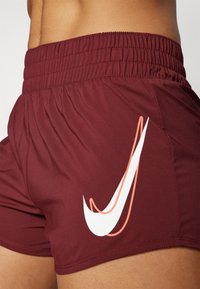 Bordové sportovní šortky s elastickým páskem, na boku s bílým logem Nike orámovaným růžovou barvou. Hladká, lehká tkanina.
