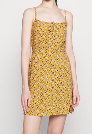 Gelbes Blumen-Sundress mit dünnen Trägern, einer Schleifendetail am Ausschnitt und einer lockeren Passform, ausgestattet mit einem seitlichen Schlitz und kleinen Gänseblümchen-Mustern.