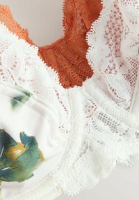 Ziedu raksta mežģīņu krūšturis, izgatavots no mīksta auduma, ar oranžiem mežģīņu akcentiem un baltiem mežģīņu elementiem, izceļot sarežģītu detalizāciju un tekstūras.