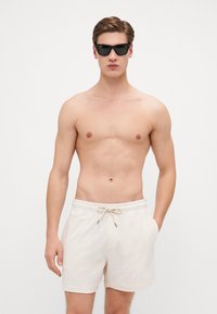Shorts en coton beige clair avec une taille élastique et un cordon de serrage, dotés de poches latérales. Modèle portant des lunettes de soleil noires, sans chemise.
