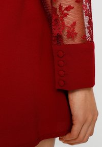 Červené šaty s krajkovými rukávy a květinovým vzorem, se čtyřmi knoflíky na manžetách. Pevná látka s hladkou strukturou.