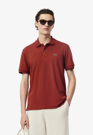 Polo shirt rossa con colletto classico, maniche corte e logo di un coccodrillo verde sul petto. Tessuto liscio con vestibilità rilassata.
