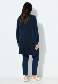 Tummansininen neulecardigan, jossa on pitkät hihat ja avonainen kaula-aukko, yhdistetty tummansinihin housuihin ja beigeihin lenkkareihin, nähtynä takaa.