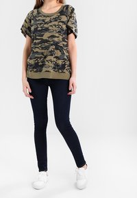 Kamuflage-mønstret olivengrøn t-shirt med korte ærmer og rund hals, parret med mørkeblå slim-fit jeans og hvide sneakers.