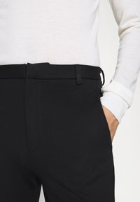 Pantalones negros de textura lisa, con frente plano y trabillas para cinturón, combinados con un suéter blanco con puños acanalados.