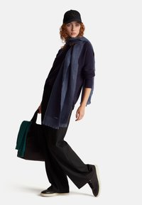 Navy knitted sweater layered with a blue fringed scarf, black wide-leg trousers, and black sneakers, carrying a black tote with a green scarf.