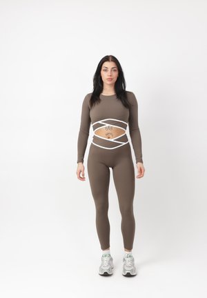 Donna in piedi, rivolta in avanti, che indossa un completo da allenamento marrone aderente con ritagli incrociati in vita e scarpe da ginnastica bianche su sfondo bianco.