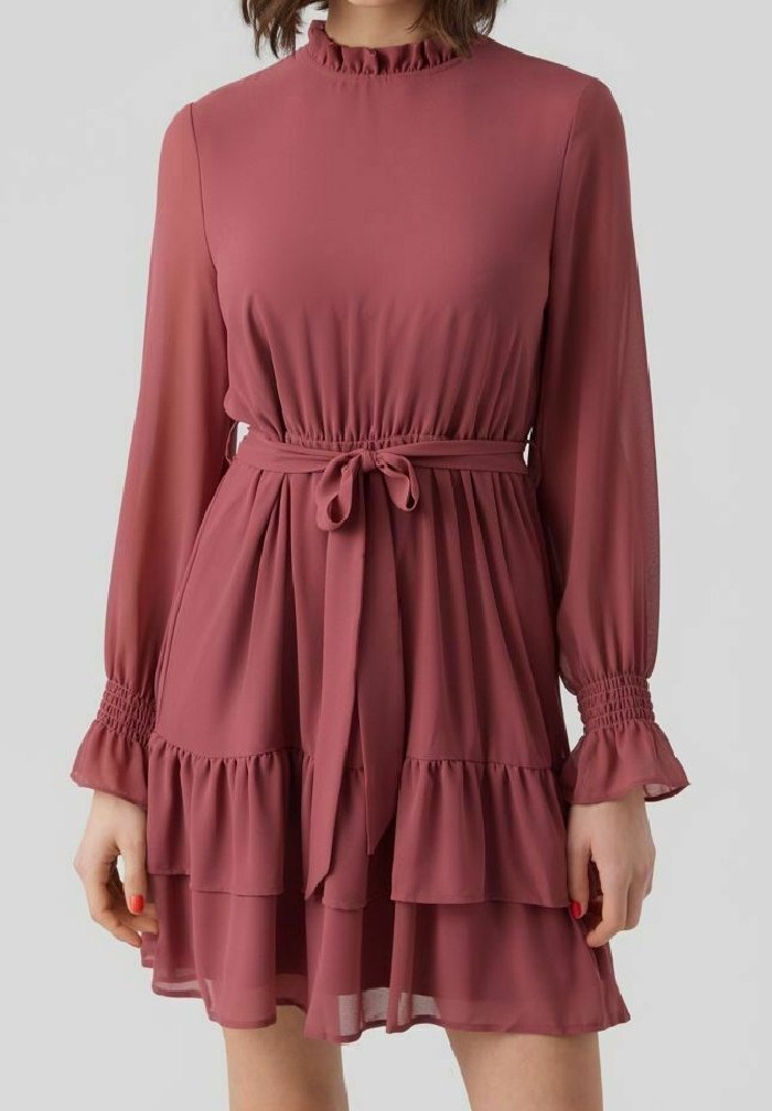 Robe à manches longues rose poudré avec col à volants, poignets smockés, ceinture à la taille et jupe à volants portée par une personne debout.