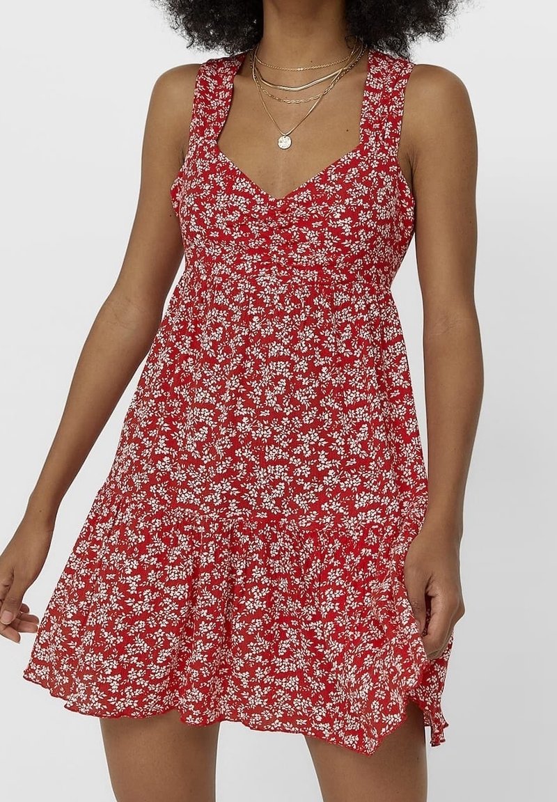 Femme portant une robe rouge sans manches à motif floral blanc et des colliers en or superposés, debout avec un bras légèrement levé.
