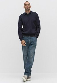 Donkerblauwe trui met een kwartrits, gedragen over een grijze T-shirt. Gecombineerd met lichtblauwe jeans en grijze sneakers. Eenvoudige, relaxte pasvorm.