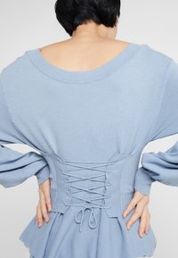 Pull bleu clair avec un large col, des manches bouffantes et un détail lacé dans le dos. Texture douce et ourlet à volants superposés qui mettent en valeur le design.