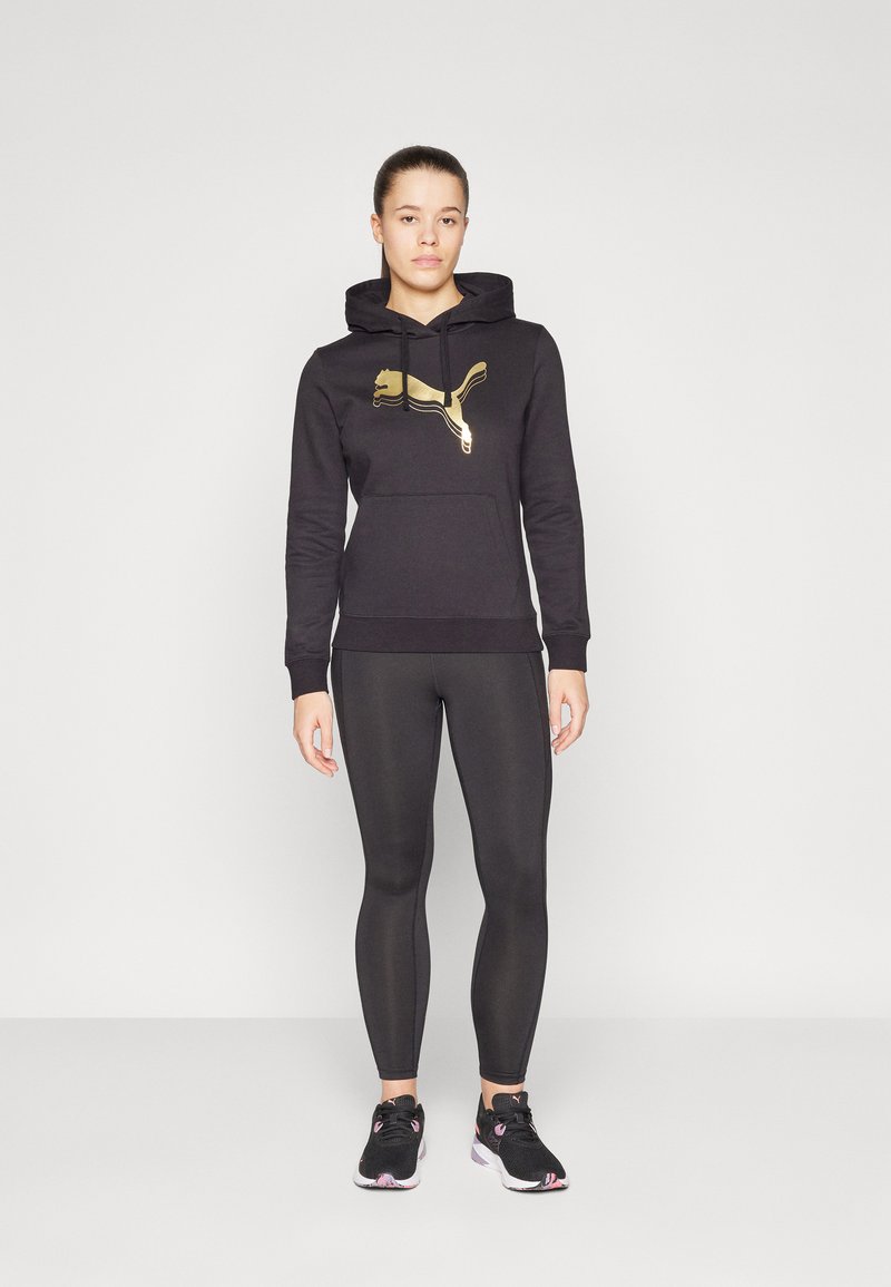 Schwarzer Hoodie mit goldfarbenem Puma-Logo, Vorderseiten-Tasche und Kordelzug. Getragen mit schwarzen Leggings und schwarzen Sportschuhen mit pinken Akzenten.