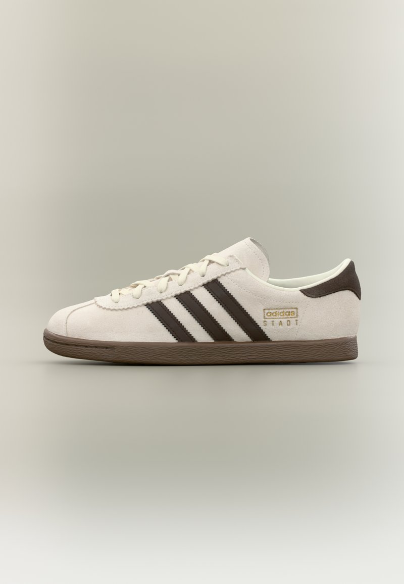 Witte suède Adidas sneaker met zwarte strepen, bruine zool en het "adidas STADT" logo aan de zijkant, opzij getoond tegen een neutrale achtergrond.