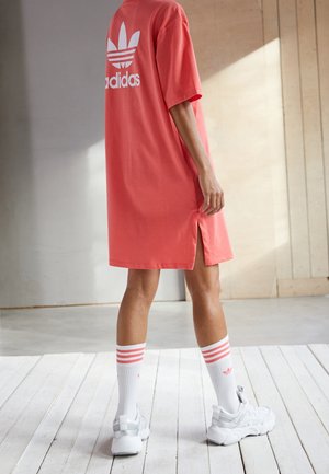 Person iført en koralfarvet Adidas-kjole, hvide Adidas-sokker med pink striber og hvide chunky sneakers, stående på hvidt trægulv.