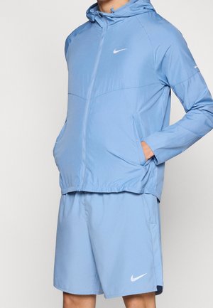 Homme portant une veste de sport Nike bleu clair avec capuche et un short assorti, mains dans les poches de la veste, sur fond blanc.