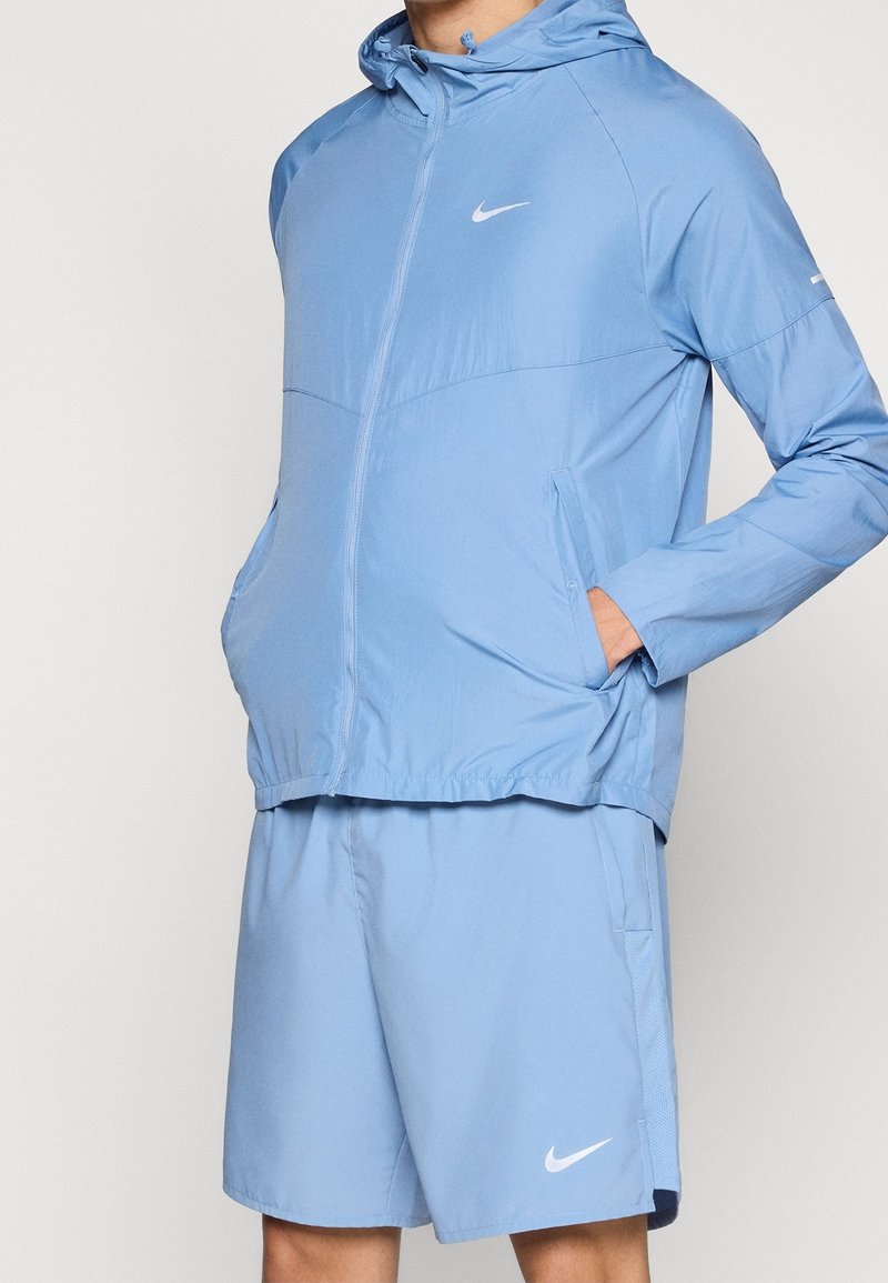 Homme portant une veste de sport Nike bleu clair avec capuche et un short assorti, mains dans les poches de la veste, sur fond blanc.