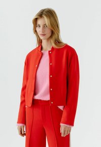 Frau trägt eine rote Jacke mit Knöpfen über einem pinkfarbenen Pullover, kombiniert mit roten Hosen, stehend vor einem schlichten hellen Hintergrund.