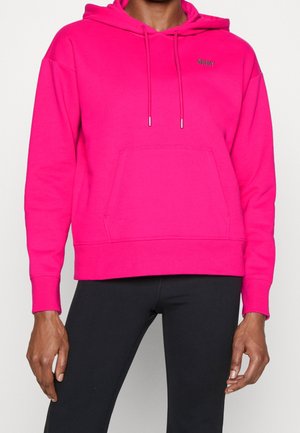 Personne portant un sweat à capuche rose vif avec poche avant et cordons, associé à un pantalon noir, visible de la poitrine aux hanches.