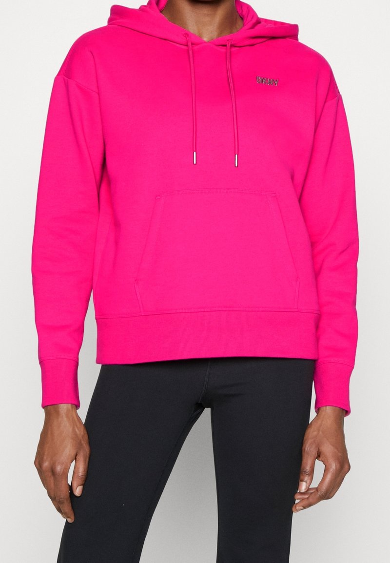 Personne portant un sweat à capuche rose vif avec poche avant et cordons, associé à un pantalon noir, visible de la poitrine aux hanches.