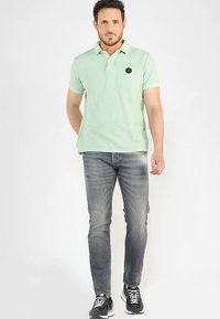 Polo de color verde claro con mangas cortas, con un logo redondo en el pecho. Combinado con vaqueros gris oscuro y zapatos deportivos negros.
