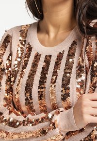 Bronze-pärlade tröja med vertikala ränder, ribbad halsringning och texturerad stickad kvalitet. Färgen är ljusrosa med reflexiga accenter.