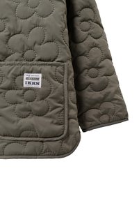 Gequiltte jas van olijfgroen materiaal, met een groot bloemenpatroon, een voorzak en een label met "IKKS HERITAGE" branding.