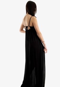 Černé maxi šaty z lehkého materiálu s tenkými ramínky a detailem na zavazování vzadu. Plynule splývají k zemi s průsvitným, lehkým vzhledem.