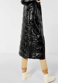 Abrigo puffer negro con acabado brillante, capucha y diseño acolchado. Presenta bolsillos laterales y cremalleras. Llevado sobre pantalones beige enrollados y botas blancas.