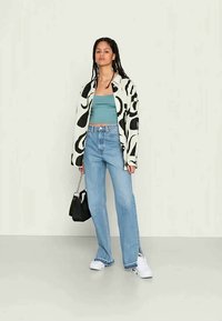 Casaco oversized com padrão preto e branco, top curto teal, jeans de perna larga em azul claro, ténis brancos e uma pequena bolsa preta.