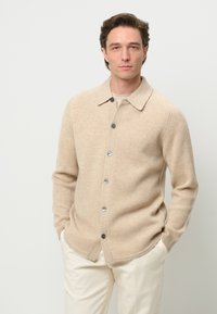 Beige ribbed cardigan with a collared neckline, front buttons, and side pockets. Textured knit fabric adds depth to the design.