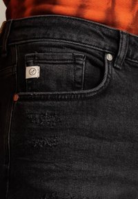 Svarta jeans i denim med en förstörd textur, silverfärgad hårdvara och en liten vävd etikett på bakfickan. Minimalistisk design.