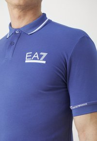Blå pikétröja med krage som har vita ränder, en logotyp "EA7" på bröstet och texten "Emporio Armani" på ärmen. Slät tygtextur.