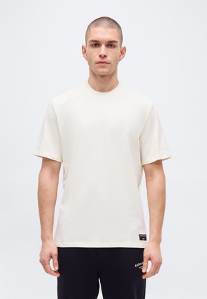 Man met kort haar, gekleed in een effen wit T-shirt met korte mouwen en zwarte broek, staand tegen een effen lichte achtergrond.