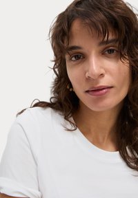 Bílý bavlněný tričko s ohrnutými rukávy, vykazuje hladkou strukturu a klasický kulatý výstřih. Modelka má kudrnaté vlasy a minimální make-up.