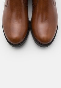 Brązowe skórzane botki sięgające do kostek o gładkiej teksturze, zaokrąglonym nosku i subtelnych detalach szycia, z czarną podeszwą i wykończeniem.