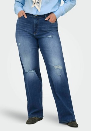 Žena v modrých opotrebovaných džínsach s rovnými nohavicami, rukami v vreckách, v svetlomodrej košeli a čiernych topánkach.