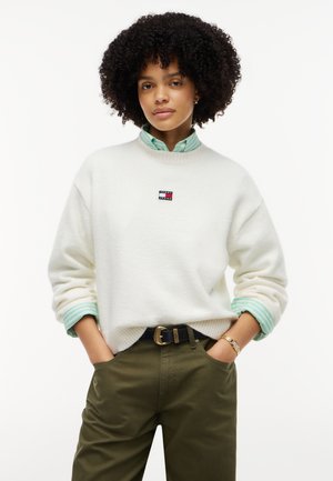 Femme aux cheveux bouclés portant un pull blanc par-dessus une chemise à rayures vertes et un pantalon vert olive, les mains dans les poches sur fond blanc.