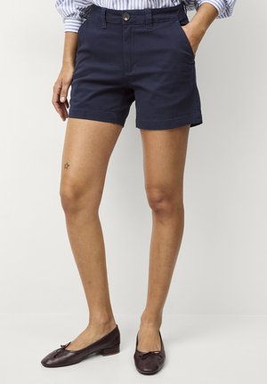 Kobieta w ciemnogranatowych szortach, włożonej w spodnie pasiastą koszuli oraz ciemnobrązowych balerinach, stojąca z jedną ręką w kieszeni.