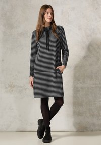 Gestreiftes graues Kleid mit langen Ärmeln, Schalkragen und Seitentaschen. Über schwarzen Leggings getragen, kombiniert mit schwarzen Stiefeletten.