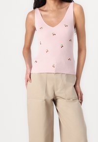 Kobieta ubrane w różowy ribbed top z małym haftem w kształcie wiśni i beżowe szerokie spodnie, pozująca z jedną ręką w kieszeni.